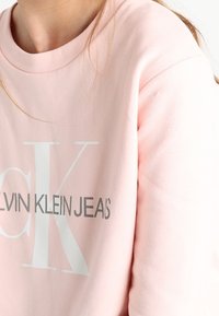 Světle růžová mikina s kulatým výstřihem, na přední straně s výrazným bílým logem "CALVIN KLEIN JEANS" v tučném písmu.