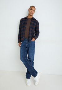 Blauwe geruite flanellen overhemd over een bruine t-shirt, gecombineerd met blauwe spijkerbroek en witte sneakers. Model poseert met één hand in de zak.
