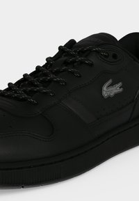 Baskets noires Lacoste en cuir avec lacets noirs et logo crocodile argenté sur le côté sur un fond uni.