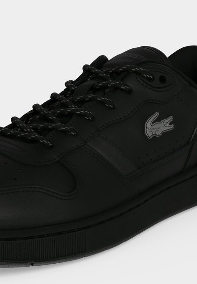 Baskets noires Lacoste en cuir avec lacets noirs et logo crocodile argenté sur le côté sur un fond uni.