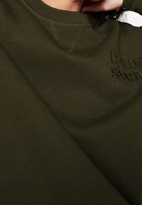 Sudadera verde oliva con textura suave, que presenta un logo bordado "LE CLUB SPORTIF" en el frente y un diseño de cuello redondo.