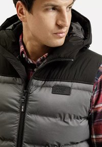 Padded vest w kolorystyce czarno-szarej, z kapturem i zamkiem. Pod nim widoczna jest koszula w kratę, a na wierzchu znajduje się metka z logo.