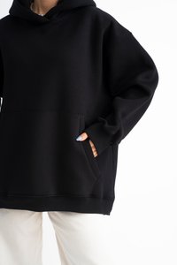 Felpa nera con cappuccio realizzata in un tessuto morbido, dotata di tasca frontale, cappuccio ampio e polsini a costine. Abbinata a pantaloni chiari.