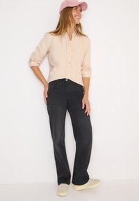 Camicia button-up beige chiaro con maniche lunghe, abbinata a jeans neri a gamba dritta e sneakers crema, su uno sfondo bianco.