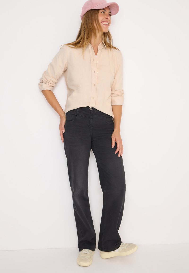 Camicia button-up beige chiaro con maniche lunghe, abbinata a jeans neri a gamba dritta e sneakers crema, su uno sfondo bianco.