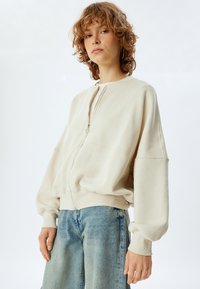 Sudadera de cremallera en un tono beige claro con hombros caídos, dobladillo y puños acanalados. Combina con unos vaqueros rectos de color azul claro. Textura suave.