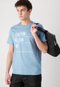 Šviesiai mėlynas medvilninis marškinėliai su balta užrašu, kuriame parašyta "CALVIN KLEIN NY" ir įkūrimo data. Modelis taip pat laiko juodą užtrauktuku uždaromą maišelį.