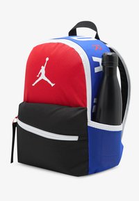 Σακίδιο πλάτης με κόκκινες, μπλε και μαύρες υφασμάτινες ενότητες. Λευκό λογότυπο εμφανές, με πλαϊνή τσέπη που κρατά ένα μαύρο μπουκάλι νερού.