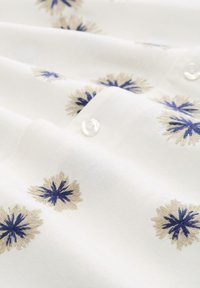 Weißer Stoff mit blauer und beiger floralem Stickerei; umfasst kleine, klare Knöpfe entlang der Kante. Glatte Textur mit detaillierten Mustern.