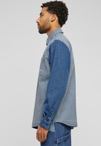 Denim-paita, jossa on yhdistelmä tummansinistä ja pystysuoria sinisiä raitoja. Varustettu kauluksella, napitettavilla hihansuilla ja rintataskulla.
