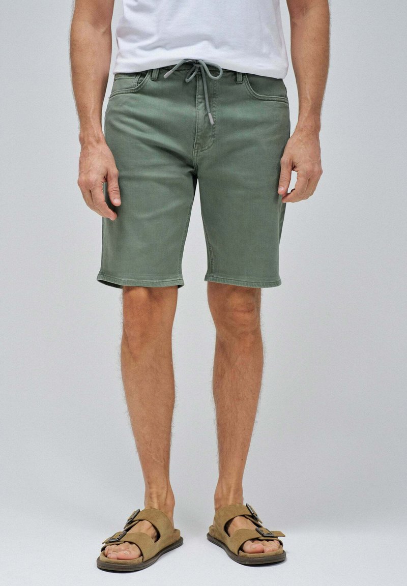 Pantalones cortos de color verde claro, hechos de una tela suave con una cintura elástica ajustable, que llegan a mitad del muslo, combinados con sandalias marrones con una hebilla decorativa.