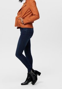 Pomarańczowa skórzana kurtka motocyklowa z pikowanymi detalami na ramionach, niebieskie dopasowane jeansy oraz czarne lakierowane botki za kostkę na grubych obcasach.