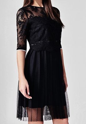 Vrouw draagt een zwarte jurk tot op de knie met een kanten lijfje, doorschijnende kanten halve mouwen en een geplooide rok.
