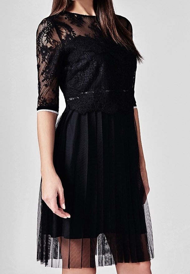 Femme portant une robe noire mi-longue avec un corsage en dentelle, des manches mi-longues en dentelle transparente et une jupe plissée.