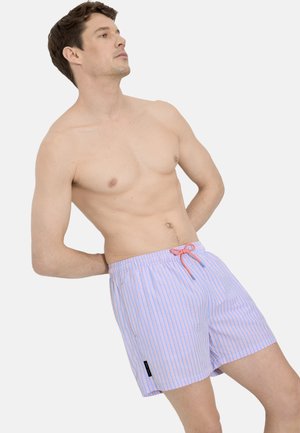 Hombre sin camiseta reclinado hacia atrás, vistiendo pantalones cortos de baño azul claro con finas rayas verticales blancas y rosas y un cordón rosa.