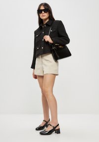 Chaqueta corta negra texturizada con botones dorados y bolsillos, pantalones cortos beige y zapatos de charol negros con tacón cuadrado. Incluye un bolso negro.
