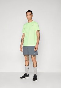 Neongrønn T-skjorte med korte ermer og logo, grå shorts og svarte joggesko. Personen har synlige tatoveringer og grå sokker.