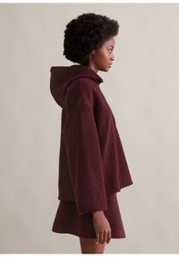 Burgundowy pulower z kapturem, wykonany z teksturowanej tkaniny. Charakteryzuje się prostym krojem i szerokimi rękawami, w komplecie z pasującą mini spódnicą.