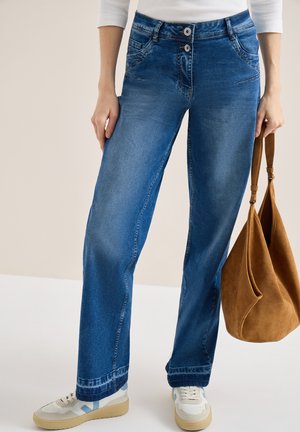Femme portant un jean large bleu et des baskets blanches, tenant un sac à main en daim brun à côté d'elle.