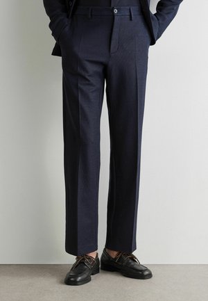 Mann trägt dunkelblaue, maßgeschneiderte Hose und schwarze Leder-Schnürschuhe, steht mit den Händen in den Taschen vor einem schlichten Hintergrund.