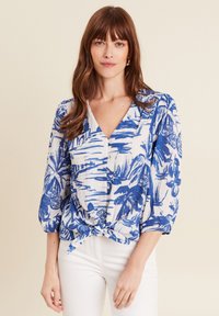 Blaue und weiße Bluse mit einem tropischen Blumenmuster, V-Ausschnitt, Knopfleiste und geknotetem Saum. Lange Puffärmel, leichtes Material.
