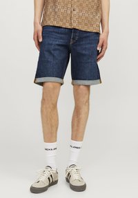 Denim šorti, tumši zili ar gludu tekstūru, ar sarullētām piedurknēm, kombinēti ar beigu krāsas sporta apaviem un baltiem zeķēm ar melnu uzrakstu.