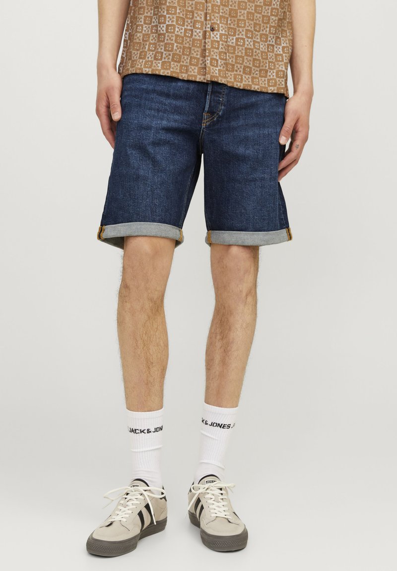 Denim šorti, tumši zili ar gludu tekstūru, ar sarullētām piedurknēm, kombinēti ar beigu krāsas sporta apaviem un baltiem zeķēm ar melnu uzrakstu.