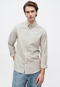 Jovem com as mangas arregaçadas, vestindo uma camisa bege clara com botões e jeans azuis, em pé contra um fundo branco simples.