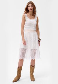 Robe en dentelle blanche avec un haut en crochet, encolure carrée et bretelles volantes ; la jupe transparente présente des motifs de dentelle complexes. Porter avec des bottes de cow-boy marron.