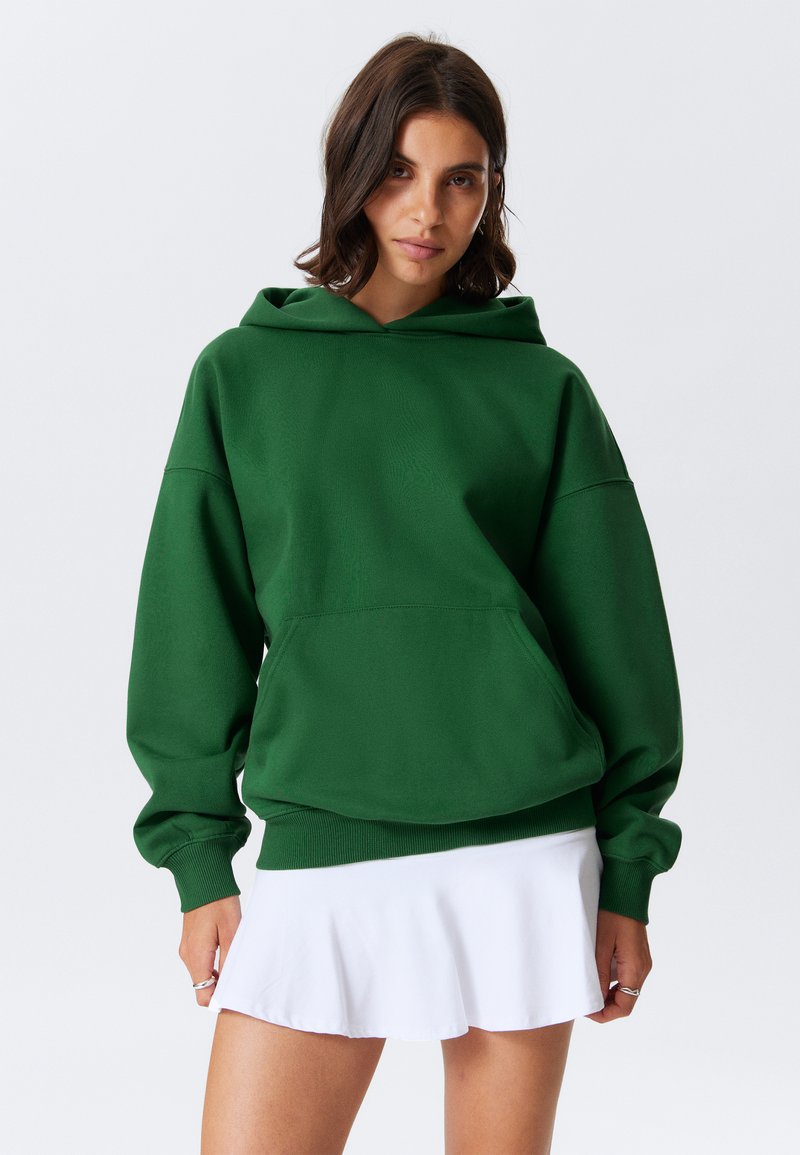Felpa verde in cotone con una grande tasca frontale, polsini a coste e cappuccio con cordini, indossata sopra una gonna bianca svasata. Design minimalista.