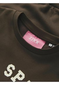T-shirt in cotone marrone con lettere bianche in grassetto che spellingano "SPORT." Include un'etichetta rosa con "JJXX" e la taglia. Collo a coste texturizzato.