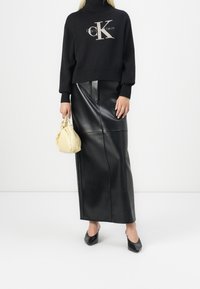 Pull noir en maille avec logo, associé à une longue jupe en cuir noire. Un petit sac pochette crème et des chaussures noires à bout pointu complètent le look.