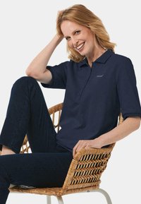 Marineblaue Polo-Shirt mit kurzen Ärmeln, ausgestattet mit einer Knopfleiste und einem kleinen Logo. Modell sitzt auf einem geflochtenen Stuhl und trägt dunkle Jeans.