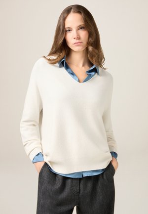 Kremowy sweter w serek o miękkiej fakturze, w zestawie z jasnoniebieską koszulą z kołnierzykiem pod spodem oraz ciemnoszarymi spodniami. Luźny strój warstwowy.