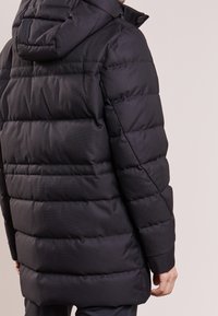 Svart pufferjacka med huva, med ett kviltat mönster och ribbade manschetter. Tyget verkar slätt och något glänsande.