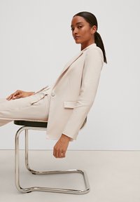 Světlobéžový přizpůsobený blazer s jedním knoflíkem a bočními kapsami, nošený s odpovídajícími kalhotami; model sedí na chromové židli.