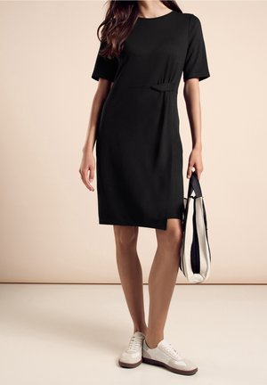 Nainen, joka pukeutunut polvipituiseen mustaan mekkoon, jossa on lyhyet hihat. Hän kantaa valkoista ja mustaraidallista kangaskassia ja käyttää valkoisia tennareita.