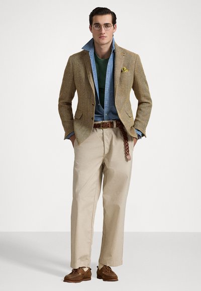 Blazer en tweed marron porté sur une chemise bleu clair, avec une chemise verte en dessous, un pantalon beige et des chaussures marron. Ceinture tressée remarquable et pochette.