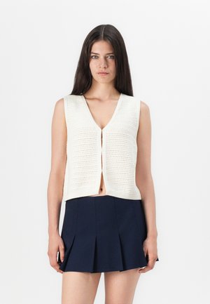 Cream knitted sleeveless vest with a V-neck, front button closure, and open design at the bottom, paired with a navy pleated skirt.
