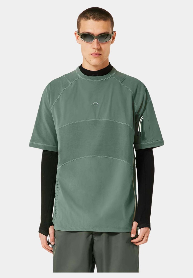 Chemise verte clair à manches courtes avec des coutures contrastées, en tissu évacuant l'humidité, et un logo sur la poitrine ; portée par-dessus une chemise à manches longues noire.