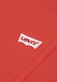Țesătură din bumbac roșu cu un finisaj texturat, având un etichetă brodată albă cu logo-ul Levi's pozicionată în colțul de jos.