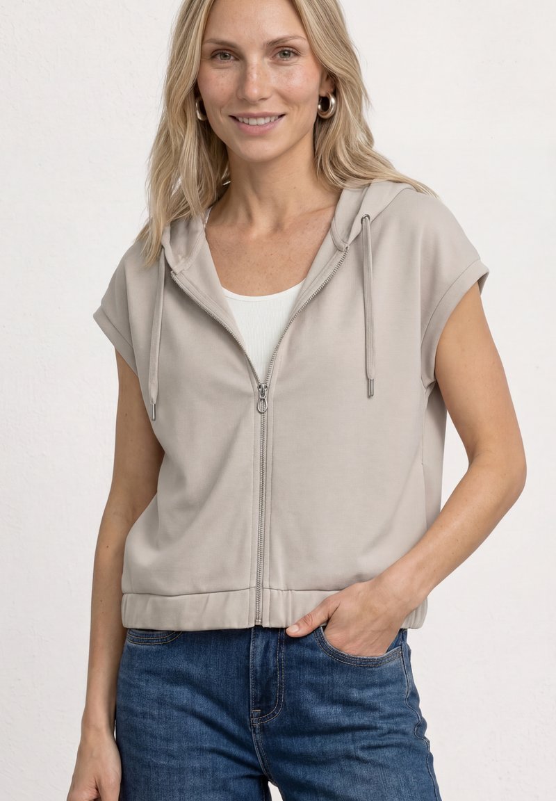 Femme souriante en sweat à capuche zippé sans manches beige clair sur un top blanc, main dans la poche d'un jean bleu, sur fond neutre.