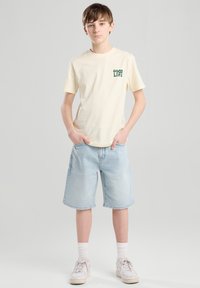 T-shirt en coton beige avec "GOOD LIFE" imprimé en vert, associé à un short en denim bleu clair et des baskets blanches à semelles texturées.