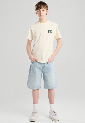 Camiseta de algodón beige con "GOOD LIFE" impreso en verde, combinada con pantalones cortos de mezclilla azul claro y zapatillas blancas con suelas texturizadas.