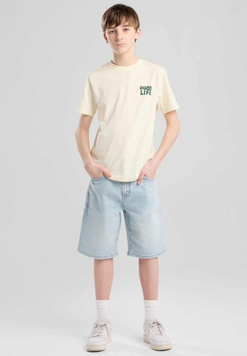 T-shirt en coton beige avec "GOOD LIFE" imprimé en vert, associé à un short en denim bleu clair et des baskets blanches à semelles texturées.