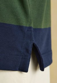 Záber zblízka na zelenú a námornícku modrú pleteninu s detailom bočného rozparku v leme, nosenú cez béžové nohavice.