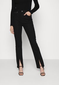 Calças flared em denim preto com fendas laterais na barra, cintura alta e design clássico de cinco bolsos. A modelo usa saltos strappy pretos.