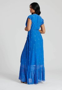Un lungo vestito blu con maniche corte a volant, in tessuto leggero, vita legata, texture con piccoli motivi lucidi e orlo a balze stratificate.