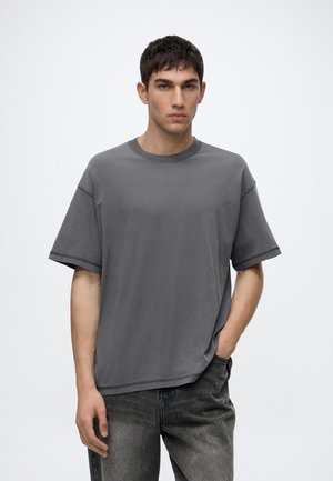 Junger Mann mit kurzen dunklen Haaren, trägt ein lockeres graues T-Shirt und dunkel gewaschene Jeans, steht mit einer Hand in der Hosentasche vor einem schlichten Hintergrund.