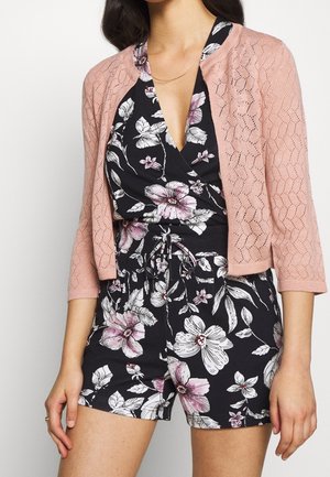 Hellrosa gestrickte Strickjacke mit Spitzenmuster-Overlay, getragen über einem schwarzen Blumen-Overall mit weißen und rosa Blumen und einem V-Ausschnitt-Design.